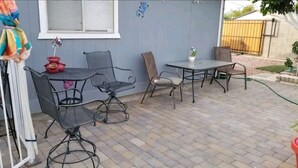 Eine Terrasse im Freien mit Tisch und Stühlen, einer kleinen Zierpflanze und einem Fenster mit Jalousien.