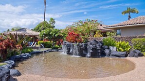 Un jardín ajardinado con un pequeño estanque, palmeras y una estructura con techo de paja.