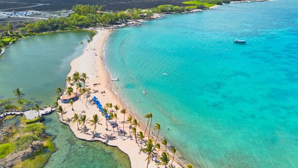 Vista aérea de una playa con aguas turquesas cristalinas, una orilla arenosa bordeada de palmeras y algunas embarcaciones.