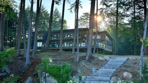 A modern cabin with a wooden exterior, surrounded by tall trees and a stone staircase leading to a deck.