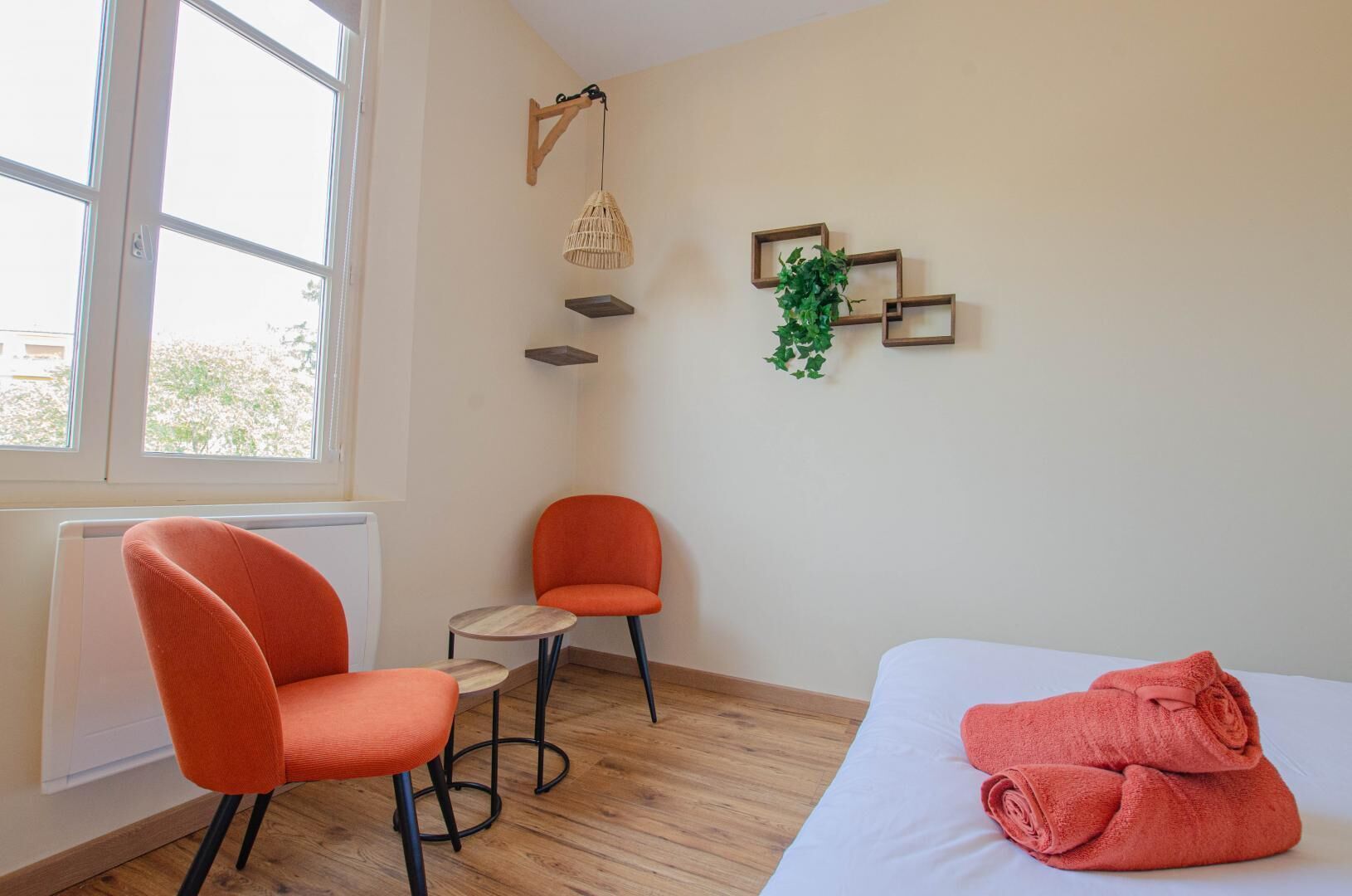 A room with a bed, two orange chairs, a small wooden table, a hanging light fixture, and a wall-mounted plant.
