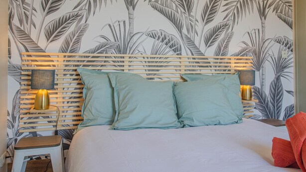 A bed with a wooden headboard, two pillows, a nightstand with a lamp, and a wall with a leaf pattern. - L'Idéal - SAINTE SAVINE (SAINTE SAVINE)