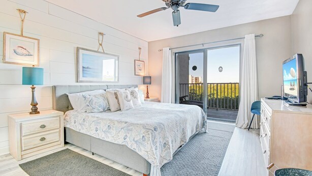 A bedroom with a bed, nightstands, a ceiling fan, a TV, and a balcony with a view.