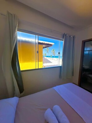 Relax in a spacious room with a large bed and a bathroom area, featuring a window with a view of the beach and palm trees.