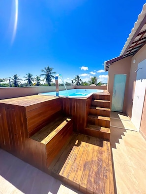 Terraza de madera con un jacuzzi y una piscina, rodeada de palmeras y un cielo despejado.
