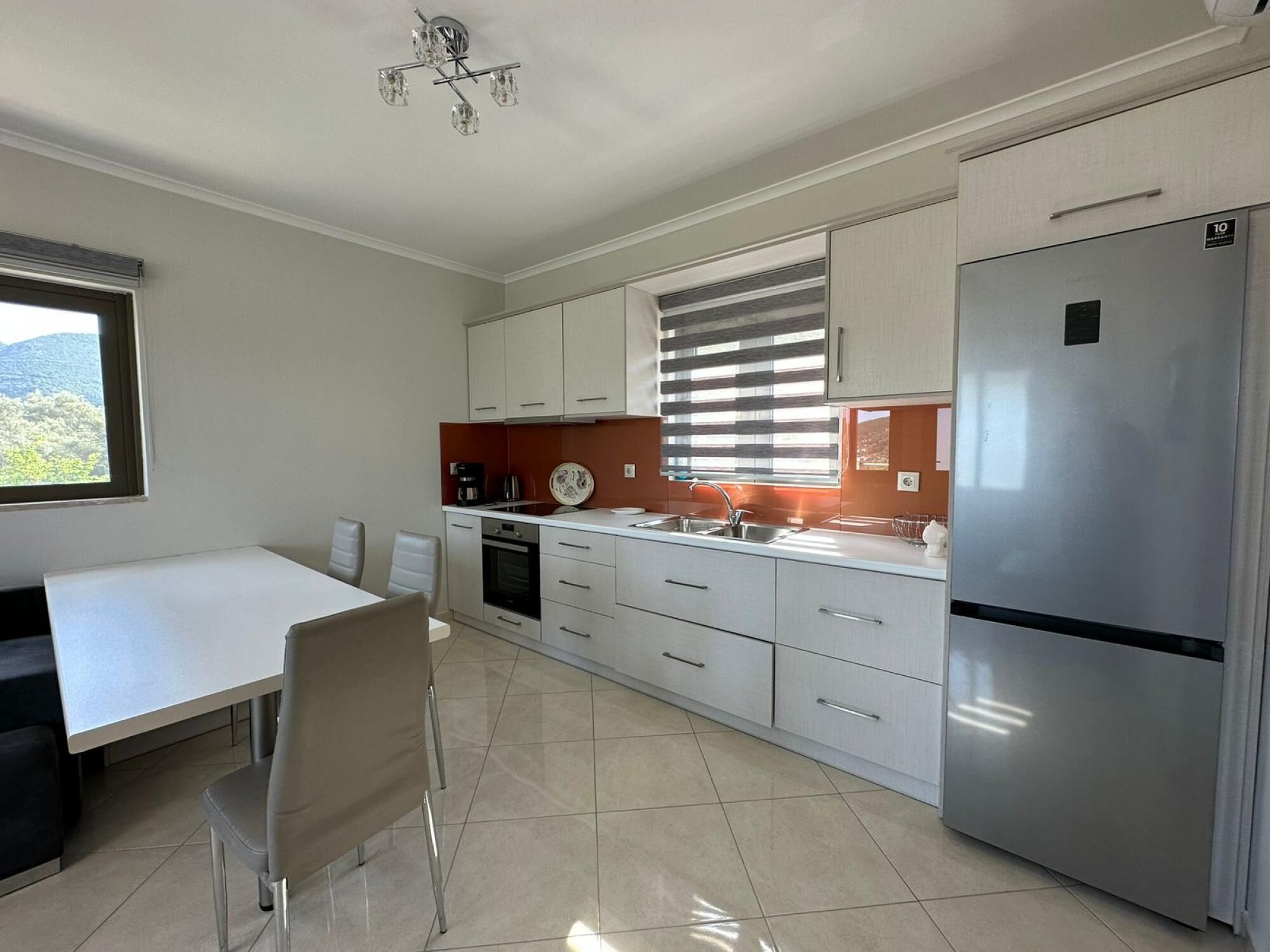 Una cocina moderna con armarios blancos, un refrigerador de acero inoxidable, una mesa de comedor con sillas y una ventana con persianas.