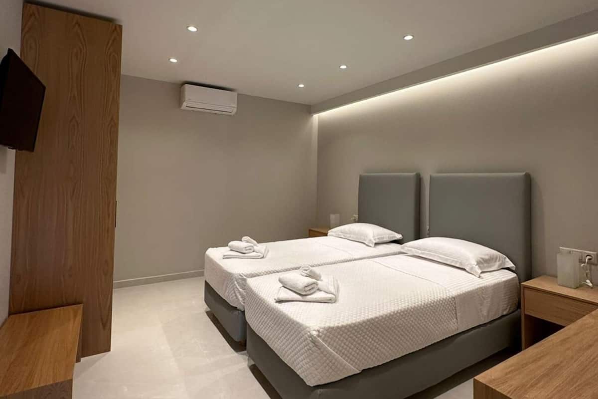 Une chambre moderne avec deux lits, une armoire en bois, un téléviseur fixé au mur et des spots encastrés au plafond.