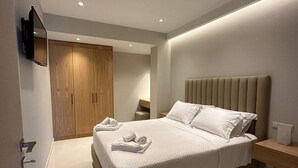 A modern bedroom with a large bed, a wooden wardrobe, a TV mounted on the wall, and a bedside table with a lamp. - Elpida Luxury Apartments & Suites (Lefkada)