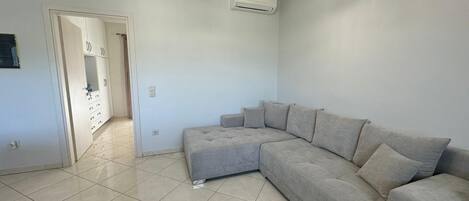 A spacious living room with a large L-shaped grey sofa, a ceiling light fixture, and a white air conditioning unit.
