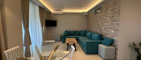 Uma sala de estar moderna com um sofá seccional, uma mesa de jantar de vidro com cadeiras, luminárias pendentes, uma TV de tela plana e uma parede decorativa com textura de pedra.