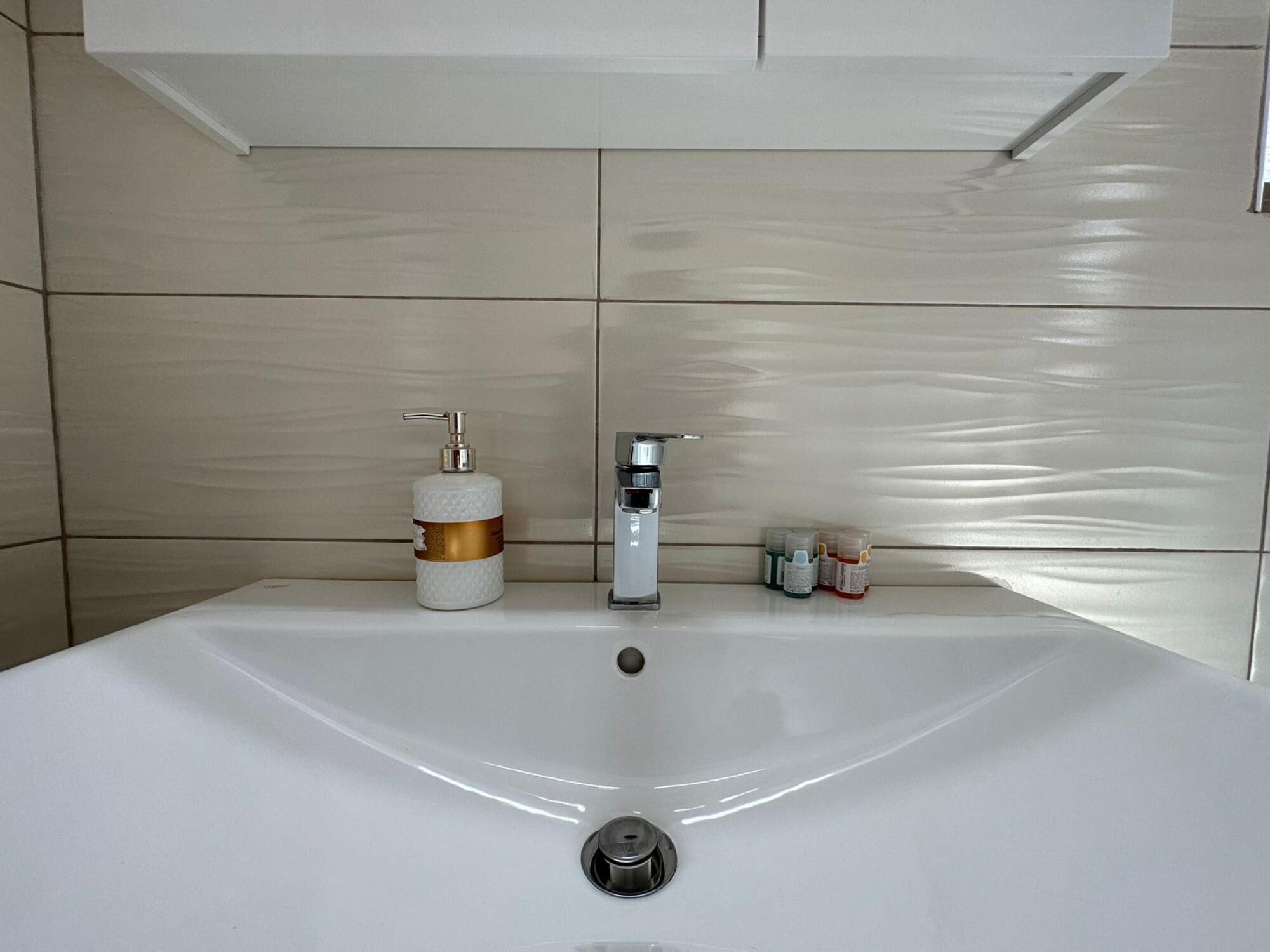 Uma casa de banho com uma pia branca, um dispensador de sabonete dourado e branco, uma torneira prateada e uma pequena garrafa de champô.