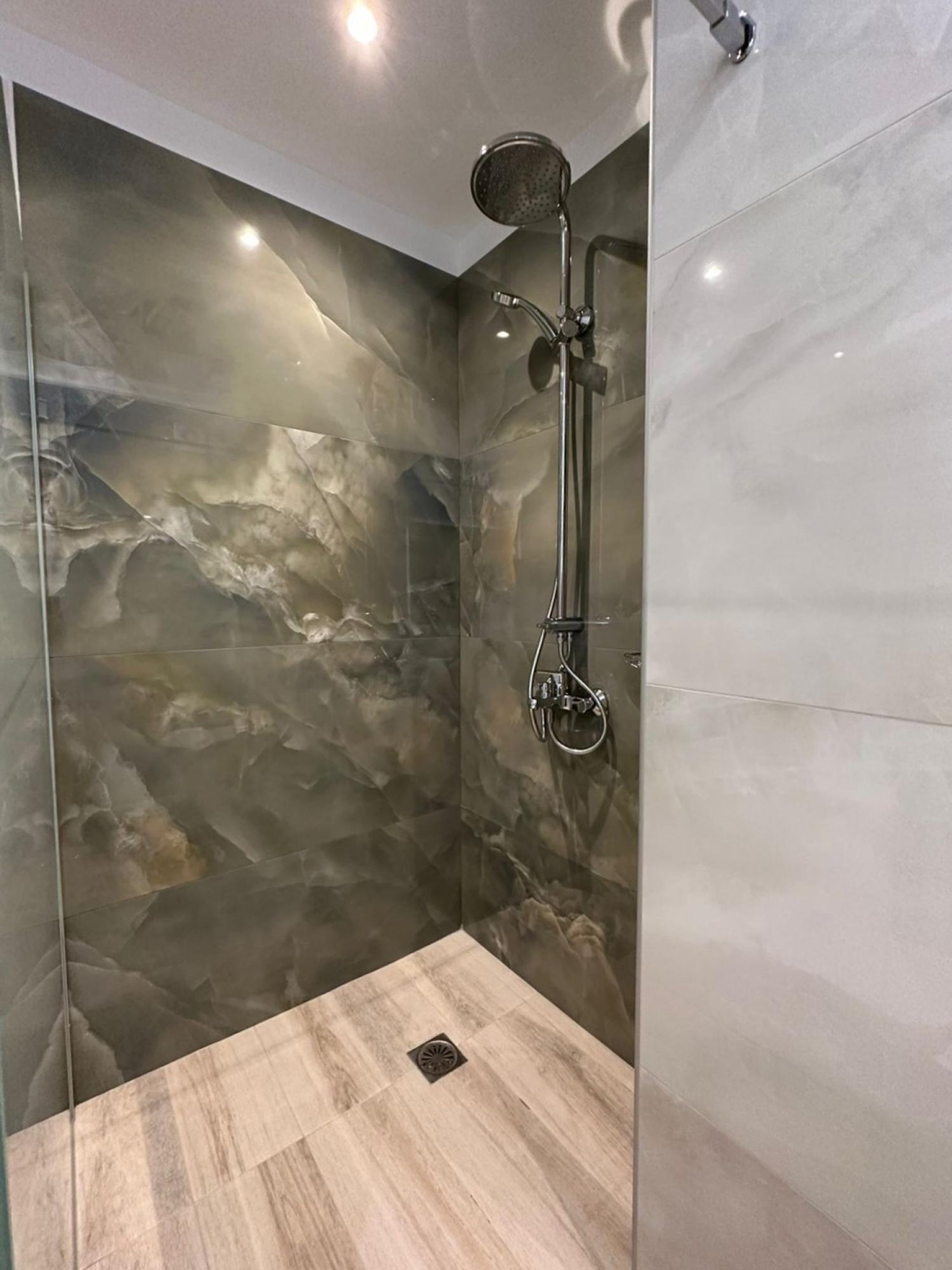 A modern shower with a glass enclosure, marble walls, and a wooden floor.