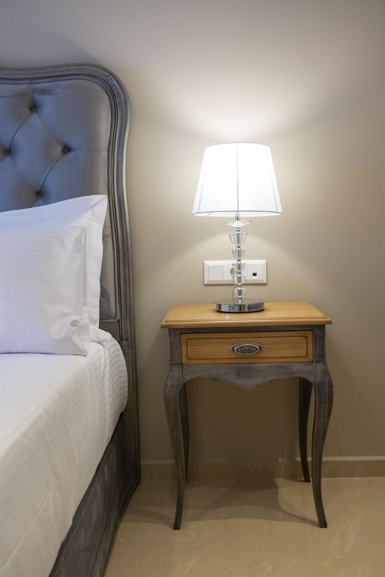 A neatly made bed with white linens, a bedside table with a lamp, and a drawer.