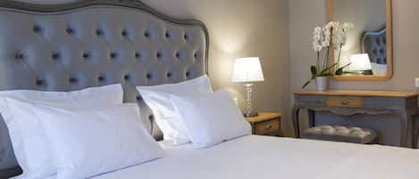 A neatly made bed with white linens and a tufted headboard. There is a bedside table with a lamp and a vase of flowers. A wooden dresser with a mirror is also visible.