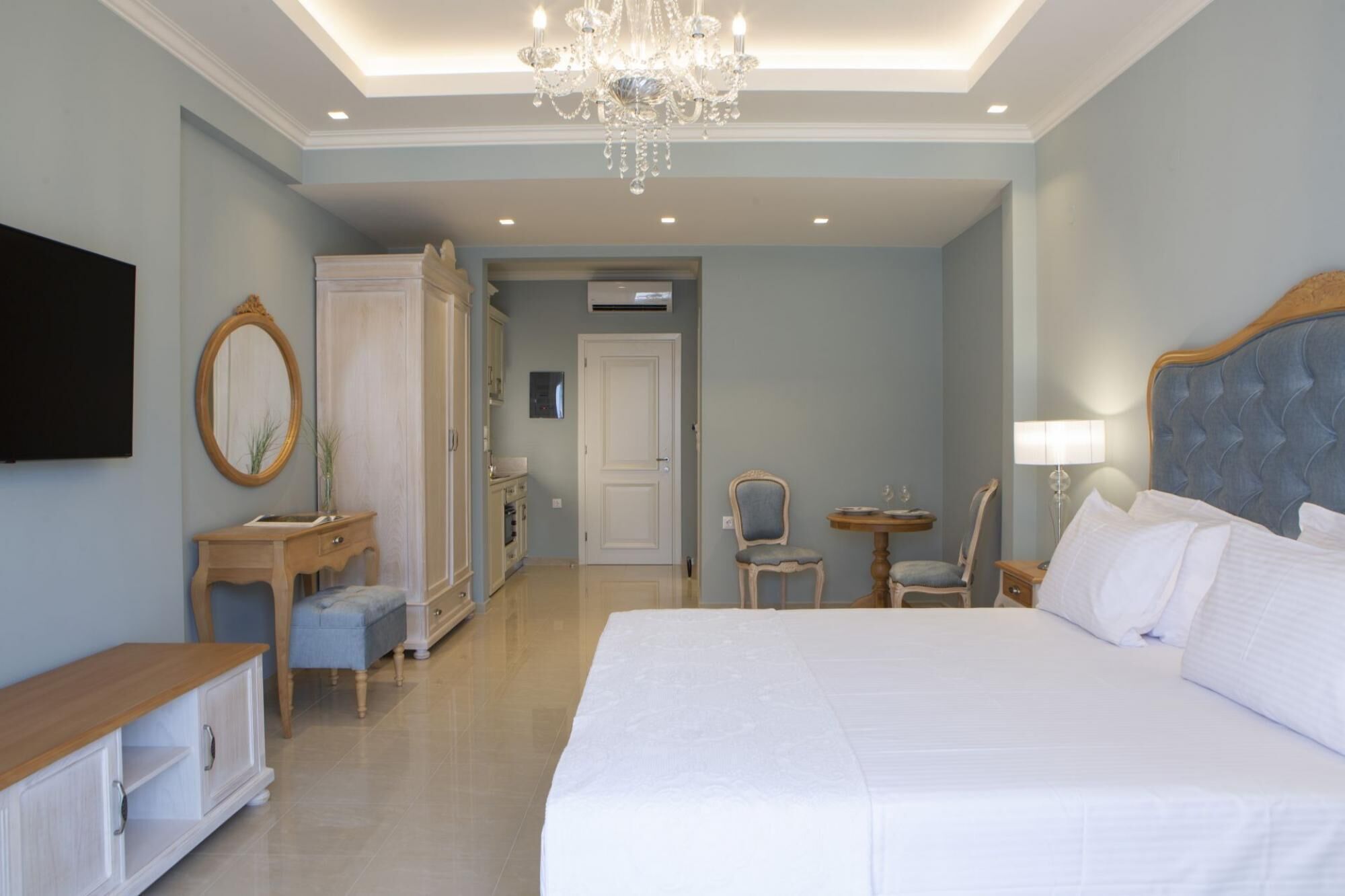 A hotel room with a large bed, a chandelier, a television, a mirror, a small table, and a chair.