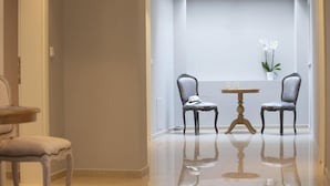 Een gang met een glanzende betegelde vloer, twee stoelen, een klein rond tafeltje en een potplant.