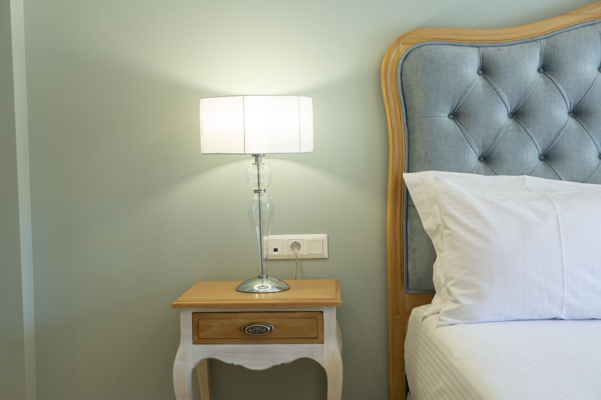 A neatly made bed with white linens, a bedside lamp on a wooden nightstand, and a wall socket.