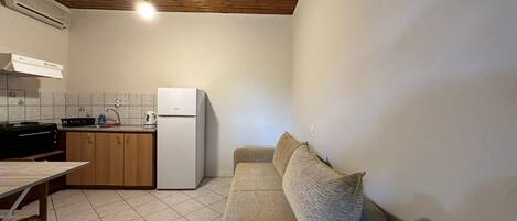 A compact kitchen with a stove, sink, and refrigerator. There is a wooden ceiling and a beige sofa with cushions.