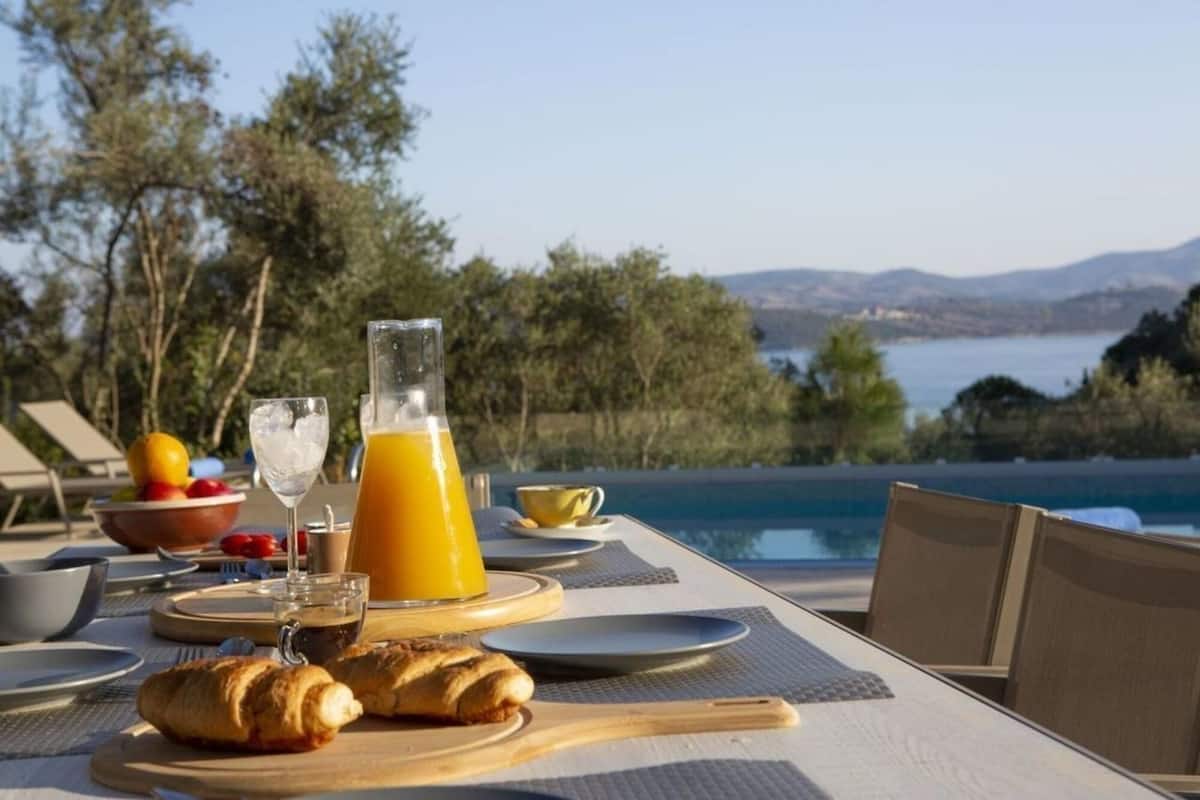 A breakfast setup with croissants, fruit, and orange juice on a table with a view of a pool and mountains.
