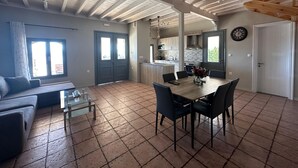 Ein großzügiger Wohnbereich mit Esstisch, Stühlen und einer Couch. Eine Küchenzeile mit Herd und Schränken ist ebenfalls vorhanden. Der Raum verfügt über eine hohe Decke mit freiliegenden Balken und ein großes Fenster.