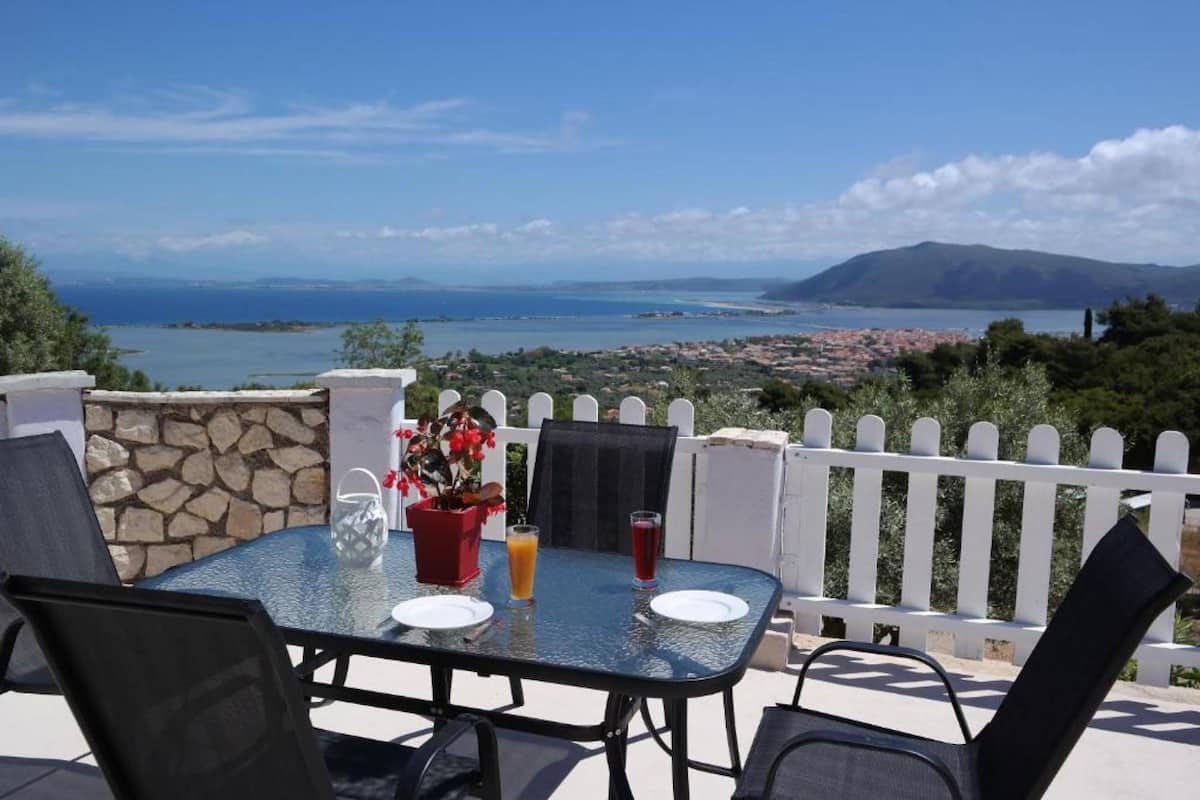 An outdoor dining area with a table and chairs, a stone wall, and a view of a coastal town and mountains.