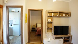 Un espacio habitable compacto con kitchenette, cama, televisor y un pequeño estante de madera.