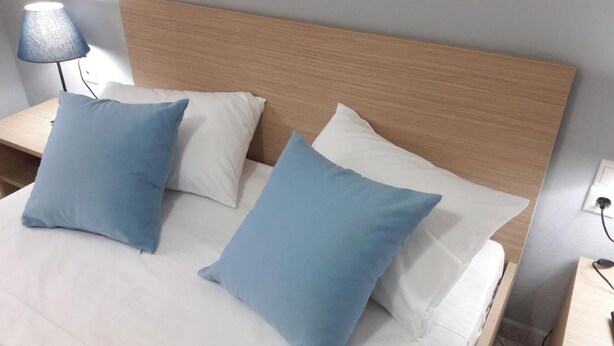 A bed with white bedding and three blue pillows, a wooden headboard, a bedside lamp, and a power outlet on the wall. - Lefkada Blue - Apartments (Lefkada)