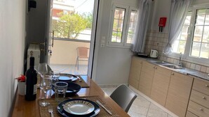 A dining area with a table set for two, a bottle of wine, and glasses. There is a window with curtains, a view of the outside, and a kitchen area with cabinets and a sink. - Lefkada Blue - Apartments (Lefkada)