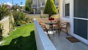 A balcony with a small table and chairs, surrounded by a well-maintained garden and a tall tree. - Lefkada Blue - Apartments (Lefkada)