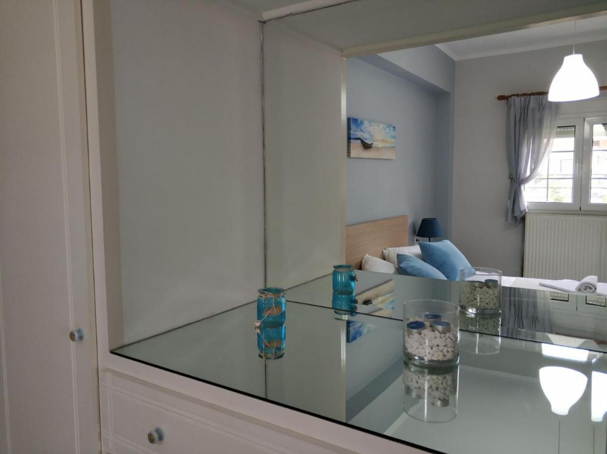 A bedroom with a glass-top table, a bed with blue pillows, a painting on the wall, and a window with curtains.