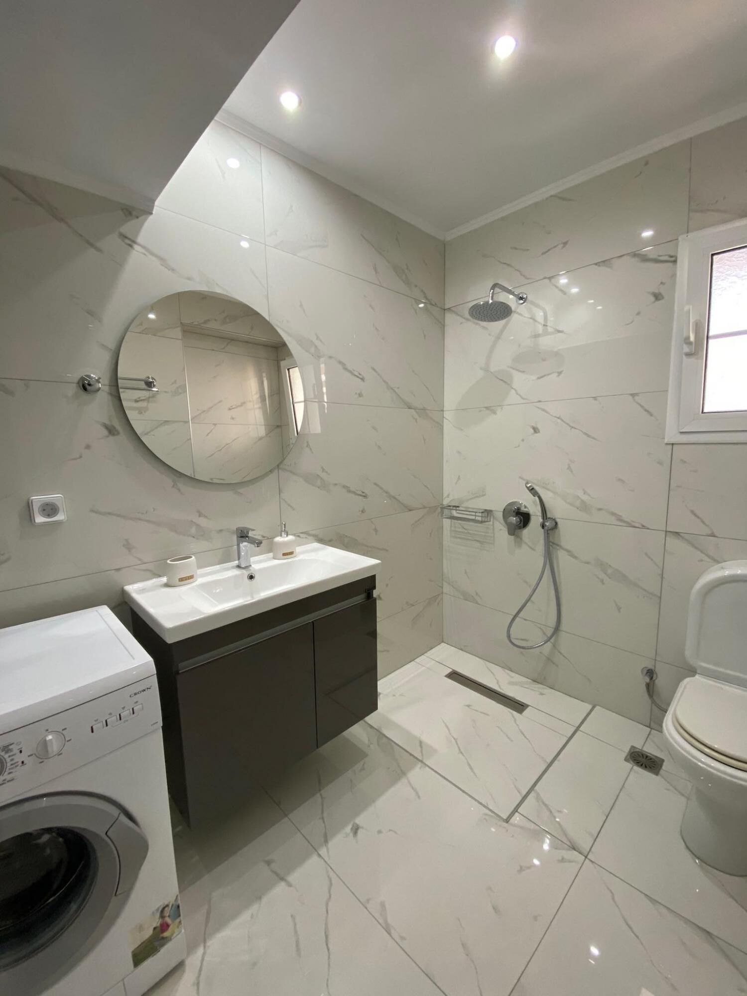 A modern bathroom with a built-in washing machine, a sink, a mirror, and a shower.