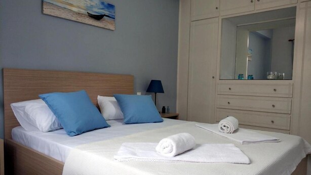 A neatly made bed with blue and white pillows, a rolled white towel, and a painting of a beach scene on the wall. - Lefkada Blue - Apartments (Lefkada)