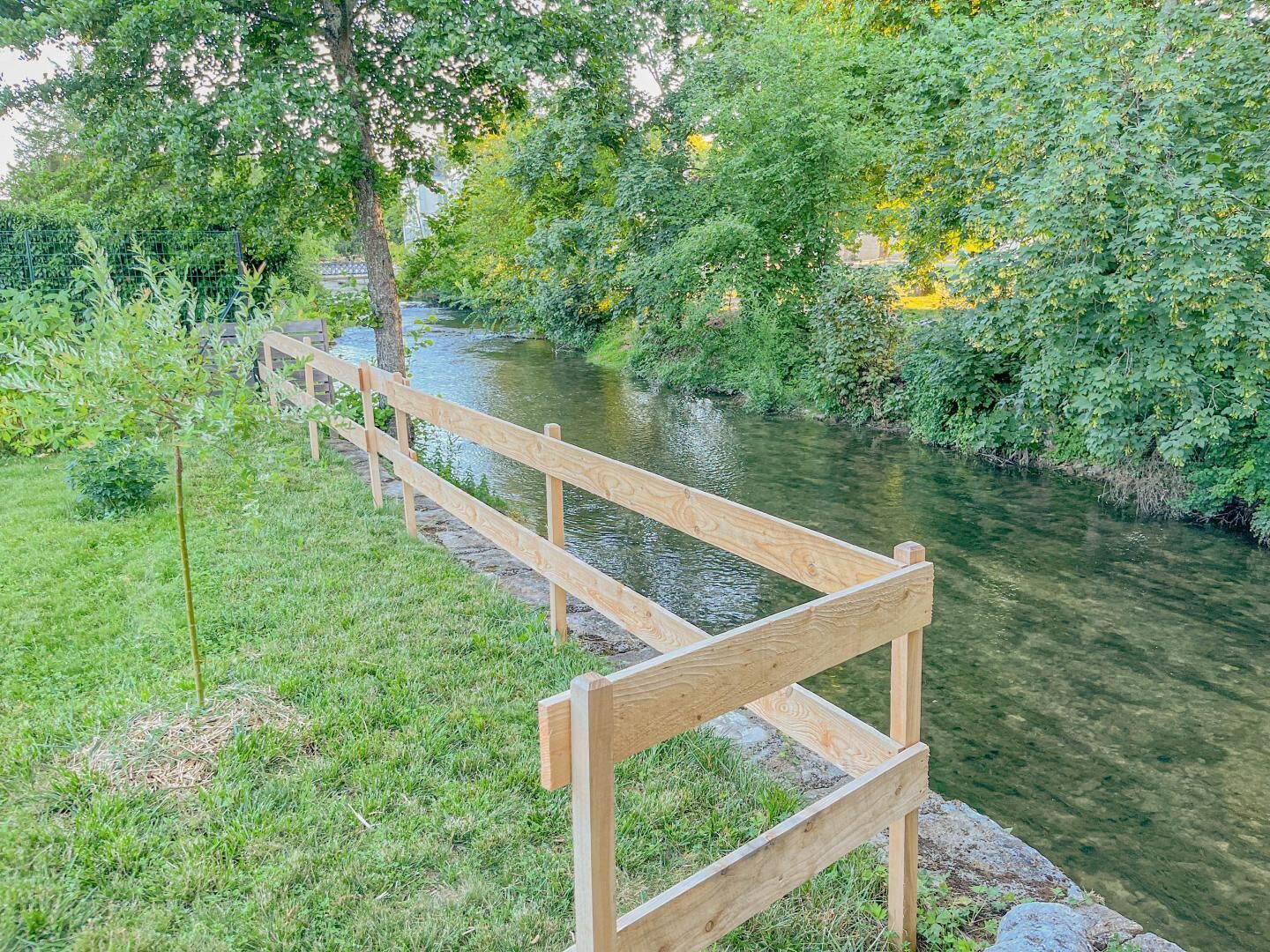 Eine Holzbank an einem Fluss, mit üppigem Grün und Bäumen im Hintergrund.