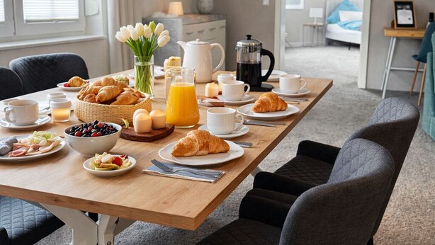 Ein Essbereich mit einem langen Holztisch, gedeckt für das Frühstück, inklusive Croissants, Früchten und Kaffee.