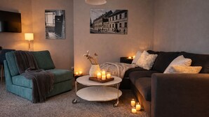 Ein gemütliches Wohnzimmer mit einem grünen Sessel, einer dunkelgrünen Couch und einem kleinen runden Tisch mit Kerzen. An der Wand befinden sich Lampen und ein Gemälde, das eine Straßenszene zeigt.