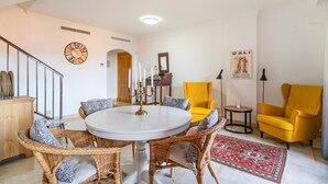 Een eetgedeelte met rieten stoelen en een ronde tafel, een gele fauteuil, een rood kamerkleed en een wandklok.