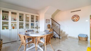 Een eetgedeelte met rieten stoelen, een witte tafel en een glazen kast met servies.