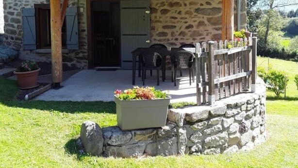 Una casa de piedra con techo de tejas, un porche de madera y un pequeño jardín con una jardinera y sillas.