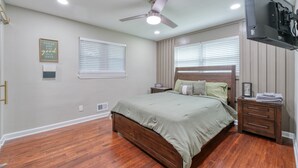 Un dormitorio con cama de madera, ventilador de techo, televisor montado en la pared y dos ventanas con persianas.