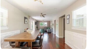 Un comedor espacioso con una larga mesa de madera y varias sillas, un ventilador de techo, y vistas a una sala de estar con un sofá y una ventana.