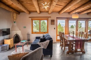 A cozy living room with a fireplace, a sofa, a dining table with chairs, and large windows offering a view of the outdoors. - Casa Lobo - Wild Relaxation - Private Hot Tub (El Prado)
