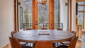 Una mesa redonda de madera con seis sillas en una habitación con amplios ventanales que dan a una zona boscosa.