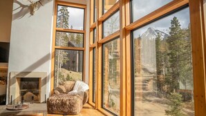 Una habitación acogedora con ventanas amplias, chimenea y vistas a un bosque y una montaña.