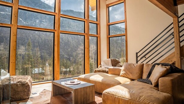 Amplia habitación con grandes ventanales que ofrecen vistas a la montaña, una escalera de madera y un sofá de cuero.