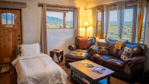 A cozy room with a bed, a leather sofa, a coffee table, and large windows offering a view of the desert landscape. - Casita de Alfarero -  A Peaceful Escape - New Hot Tub! (El Prado)
