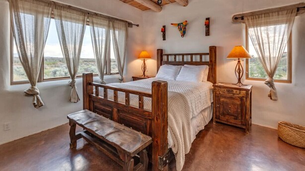 A rustic bedroom with a wooden bed, two bedside lamps, a bench, and a view of the desert through large windows. - Casita de Alfarero -  A Peaceful Escape - New Hot Tub! (El Prado)