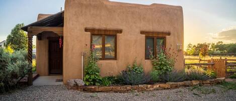 Une maison rustique en adobe, avec une porte en bois, deux fenêtres et une allée gravillonnée.