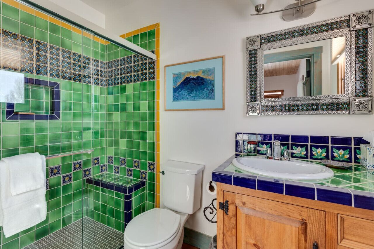 Un baño con azulejos verdes y azules, un inodoro blanco, un mueble de madera y un cuadro enmarcado en la pared.