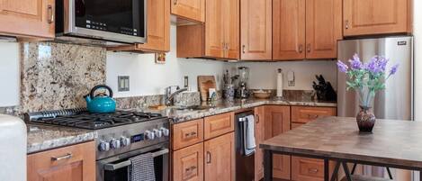 Una cocina con armarios de madera, un refrigerador de acero inoxidable, una tetera azul y un florero con flores sobre la encimera.