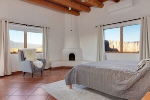 Une chambre à coucher avec un lit, un fauteuil recouvert d’un plaid en fourrure, une cheminée et de grandes fenêtres donnant sur la campagne.